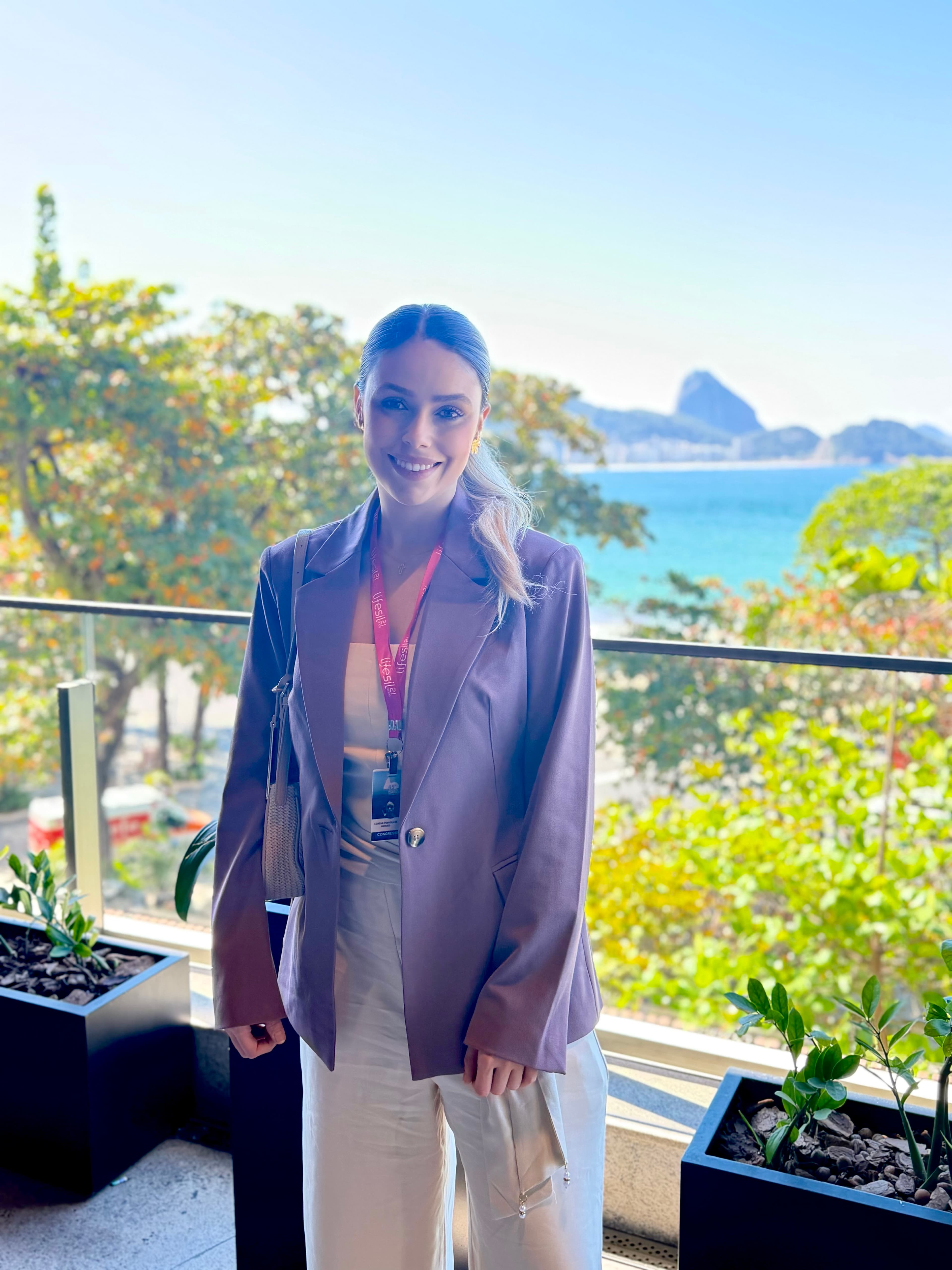 Mulher sorridente de blazer lilás em varanda com vista para o mar e montanhas cariocas.